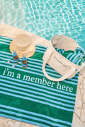 Green and blue striped towel with 'I'm a member here' text, straw hat, and bottles by a pool.