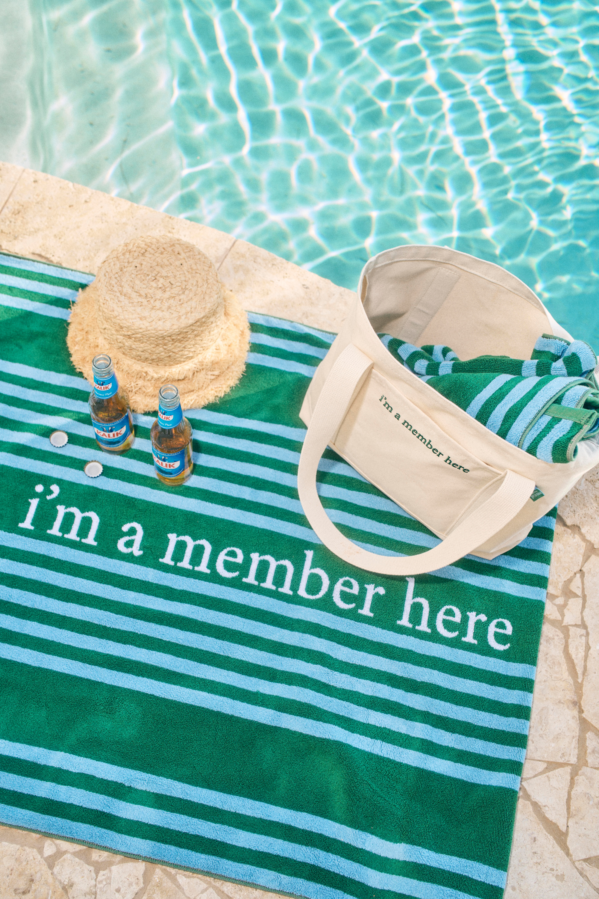 Green and blue striped towel with 'I'm a member here' text, straw hat, and bottles by a pool.